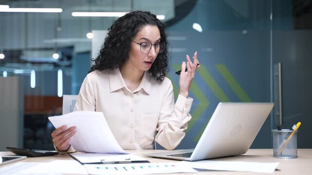 Confused businesswoman having difficulty with paper work checking documents on laptop computer while sitting at workplace in business office. Frustrated female financier unhappy with financial results