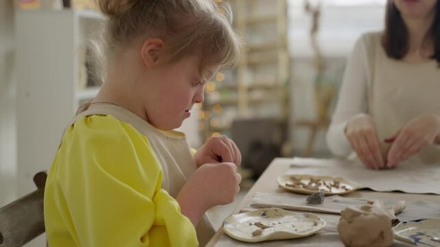 Focused girl with Down syndrome shapes clay in bright, inclusive pottery studio. Scene underscores the importance of social acceptance and belonging. Concept: social inclusion, community support