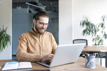 Fototapeta premium A male professional works confidently on a laptop at a wooden desk in a stylish office, focusing on business tasks with dedication.