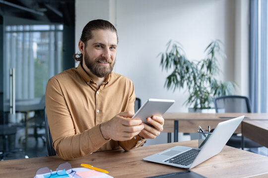 Smiling professional in casual attire working with a tablet in a modern office environment. A laptop is placed on the desk next to him, depicting a productive and professional setting. - Powered by Adobe