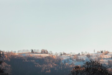 Frosty hills stretch across the horizon, bathed in warm sunlight. The terrain, adorned with patches of snow, creates a serene winter wonderland. Trees dot the landscape with a quiet charm