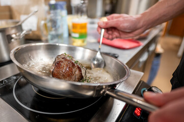 A close-up of a chef searing a piece of meat in a stainless steel skillet on a stovetop. Aromatic herbs and sizzling oil enhance the cooking process, creating a delicious atmosphere in modern kitchen.