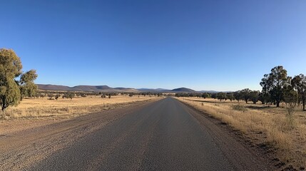 A wide-angle photo of the empty road in Australia's Outback, with red earth and sparse vegetation on both sides, a clear blue sky, and distant hills in the background.
