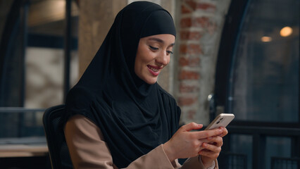 Muslim woman in hijab islamic girl Arabian businesswoman entrepreneur calm smiling business lady working at desk scrolling mobile phone gadget addicted female online app cellphone internet in office