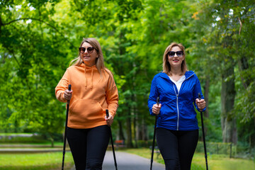 Portrait of two pretty mature women practicing Nordic walking in city park on spring day. Training...