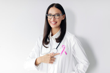 Positive woman doctor oncologist pointing at pink ribbon, standing on white studio wall background....