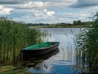 Obraz premium Serene lakeside landscape, green rowboat moored in reeds, reflective calm water, lush green trees and grass, puffy white clouds, summer day, peaceful countryside scene, photorealistic, high detail, so