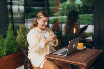 Smiling woman sitting in cafeteria holding smartphone and working on laptop. Successful businessman...