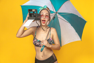 Emotional Asian girl in swimsuit with headband under umbrella with instant camera. Isolated