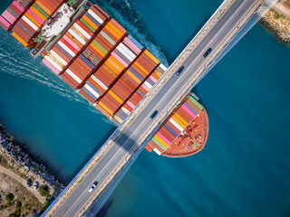 Aerial overhead closeup view of a large container cargo ship passing under a bridge with car traffic