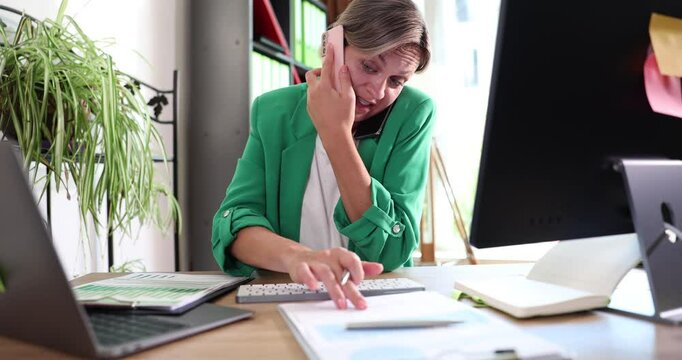 A focused woman dressed in green is coordinating multiple tasks while speaking on the phone in her home office setup