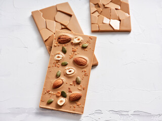 A brown chocolate bar is adorned with almonds, hazelnuts, and pumpkin seeds, resting elegantly on a light surface. Broken pieces of chocolate add visual interest