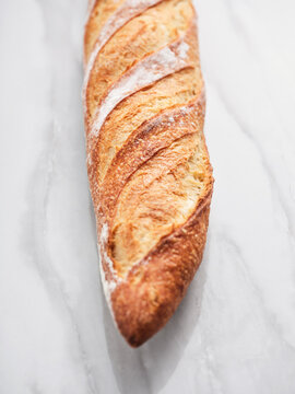 A long, crusty baguette lies on a white marble countertop. The golden-brown bread features a crispy outer texture with distinct slashes, showcasing its artisanal quality