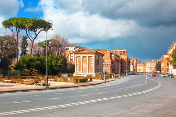 Street in Rome highlighting classical architecture with a prominent building featuring columns and a pediment, reminiscent of ancient Roman design. Ancient and modern elements alongside the street 