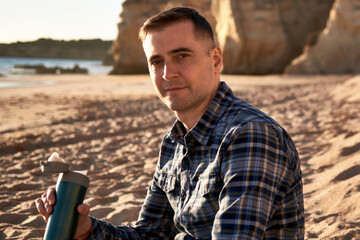 Obraz premium Portrait of a man sitting on a rocky beach during sunset and holding a thermal mug in his hand