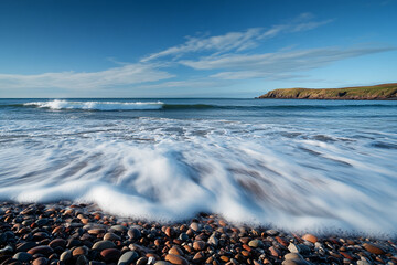 Pebbled shoreline with ocean waves and distant green cliffs under blue sky
