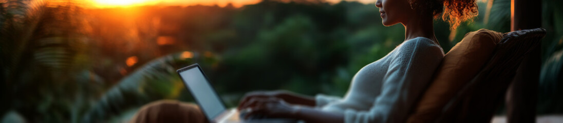 woman working on her laptop while sitting in an outdoor chair with a wooden frame, overlooking the jungle landscape at sunset. 