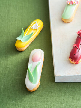 Exquisite pastries featuring vibrant colors and intricate floral designs are arranged on a textured surface. Spring themes are highlighted with egg motifs and leaf details