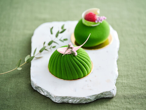 Two beautifully crafted green desserts sit on a white marble platter, decorated with fresh fruits and delicate garnishes. The desserts showcase rich textures and vibrant colors