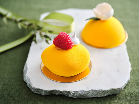 Two vibrant yellow desserts decorated with fresh raspberries and edible flowers rest on a marble plate, showcasing a modern culinary design against a green background