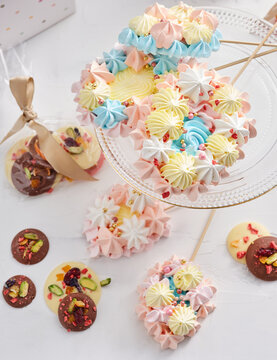 A vibrant assortment of meringue treats in pastel colors is arranged on a glass plate, surrounded by chocolate discs adorned with nuts and sprinkles, perfect for a festive gathering