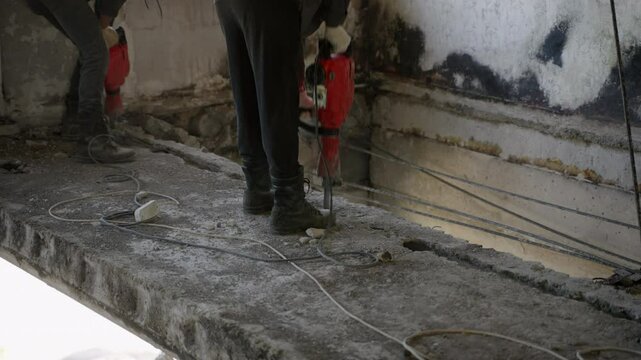 Construction workers use jackhammers to tear down damaged ceilings, walls in burned, war-affected apartment for renovation. Boots, tools, debris illustrate tough work conditions. Slow motion.