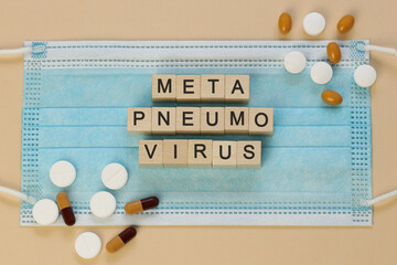 Metapneumovirus spelled with wooden cubes on a surgical mask, surrounded by pills. Blue face mask. White and brown tablets and capsules. English, German, Spanish, Italian.