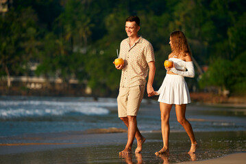Fototapeta premium Romantic couple stroll barefoot on sandy beach, man and woman in summer clothes hold hands enjoying sunset. Pair with tropical drinks walk by sea, honeymoon vacation mood on exotic island getaway.