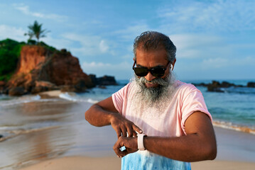 Senior Sri Lankan man with fitness tracker stands on beach. Uses smartwatch for training, wellbeing, wearing wireless earbuds. Modern tech enhances health. Ocean view, rocky shore. Healthy lifestyle.