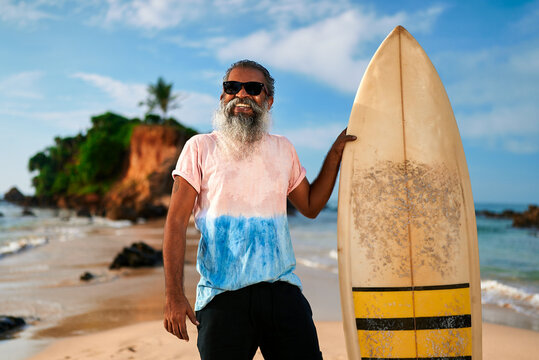 Senior man surfer stands on sandy beach with surfboard. Smiling elderly male enjoys active retirement. Ocean waves crash on shore. Surfing in happy golden years. Lifestyle, freedom, adventure.