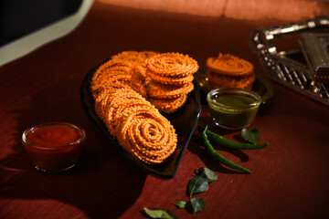 A black bean on a brown and natural background with Maharashtrian food Chakli on it along with curry leaves, green chutney, red chutney sauce It is specially made during the festival of Diwali.