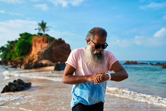 Senior man with grey beard runs on sandy beach, uses smart watch for fitness tracking. Bearded elderly male listens music in wireless earbuds. Active lifestyle tech health check.