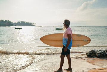 Senior man with surfboard stands on sunny beach. He looks at sea waves. Active lifestyle, surfing, ocean adventure. Ideal for travel or sports promotion. Golden hour lighting adds warmth to the scene.
