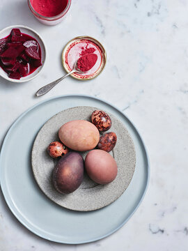 Dyed eggs in various shades sit on a gray plate. Nearby, bowls contain beetroot and a vibrant color paste, suggesting an artisanal egg-dyeing activity