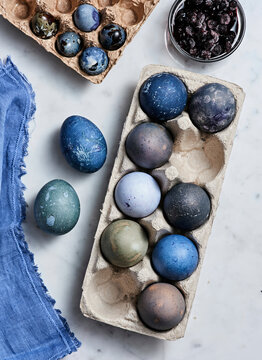 Colorful eggs rest in a carton, showcasing various hues created with natural dyes. A bowl of blueberries and a textured fabric add to the creative atmosphere of the setup