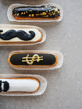 Eclairs in various shapes and colors are lined up on a grey surface, featuring playful designs like mustaches and dollar signs, perfect for a dessert table or celebration
