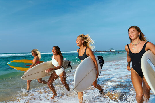 Four young women with surfboards run on sunny beach. Female surfers enjoy summer vacation at ocean shore. Friends in swimwear ready for surfing waves. Active lifestyle, beach fun.