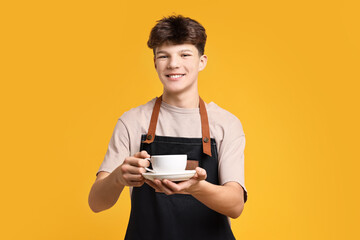 Teenage boy with cup of coffee working as barista on orange background