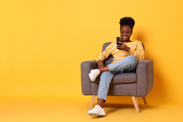 Smiling woman with smartphone sitting in armchair on yellow background. Space for text