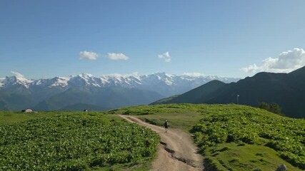 Aerial of solo backpacker hiking through alpine meadows in high mountains. Person with backpack walks along dirt path. Scenic view of green hills, snowy peaks under blue sky. Drone shot - Powered by Adobe