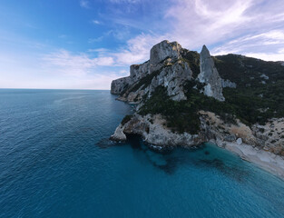 Aerial view of Punta Goloritzè along the Costa di Baunei in Sardinia, showcasing towering limestone cliffs, lush vegetation, and crystal-clear turquoise waters meeting secluded sandy coves under a vib