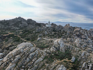 The Capo Testa lighthouse stands atop rugged granite cliffs, surrounded by wild vegetation and overlooking the vast, serene sea under a cloudy sky in Sardinia.