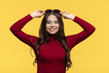 Fototapeta premium Pretty young lady in red dress and sun glasses is posing with hand on face on yellow background.