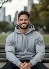 Obraz premium Portrait of a young man sitting on a wooden bench in a park. he is wearing a grey hoodie and black pants. he has a beard and is smiling at the camera.