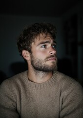 Obraz premium Close-up portrait of a young man with curly hair and a beard. he is wearing a beige sweater and is looking off to the side with a thoughtful expression on his face.