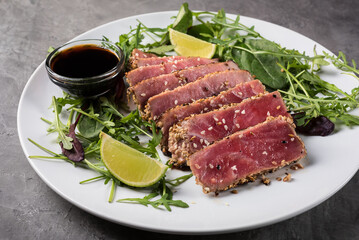 Grilled tuna steak with sesame seeds, cut into pieces with arugula and lime on a white plate on a grey background, side view.