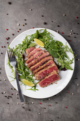 Grilled tuna steak with sesame seeds, cut into pieces with arugula and lime on a white plate on a gray background, top view.