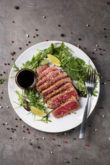 Grilled tuna steak with sesame seeds, cut into pieces with arugula and lime on a white plate on a gray background, top view.
