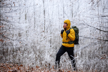 Hiking in winter forest. Hiker man in yellow jacket with backpack walking through frosty woodland