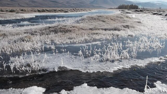 Fast-moving stream of narrow river runs along frozen banks on sunny day. Mountain landscape of autumn nature with melting ice islands and frosted grass in highland. Altai